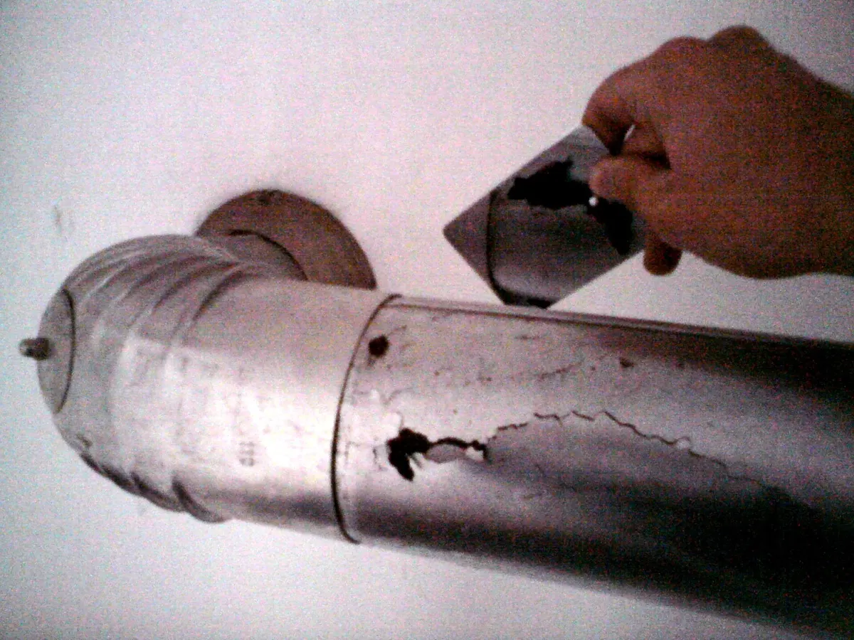 A hand holding a rusted-through piece of metal flue pipe from a gas heater next to the corroded pipe it came from, which has holes along its seam