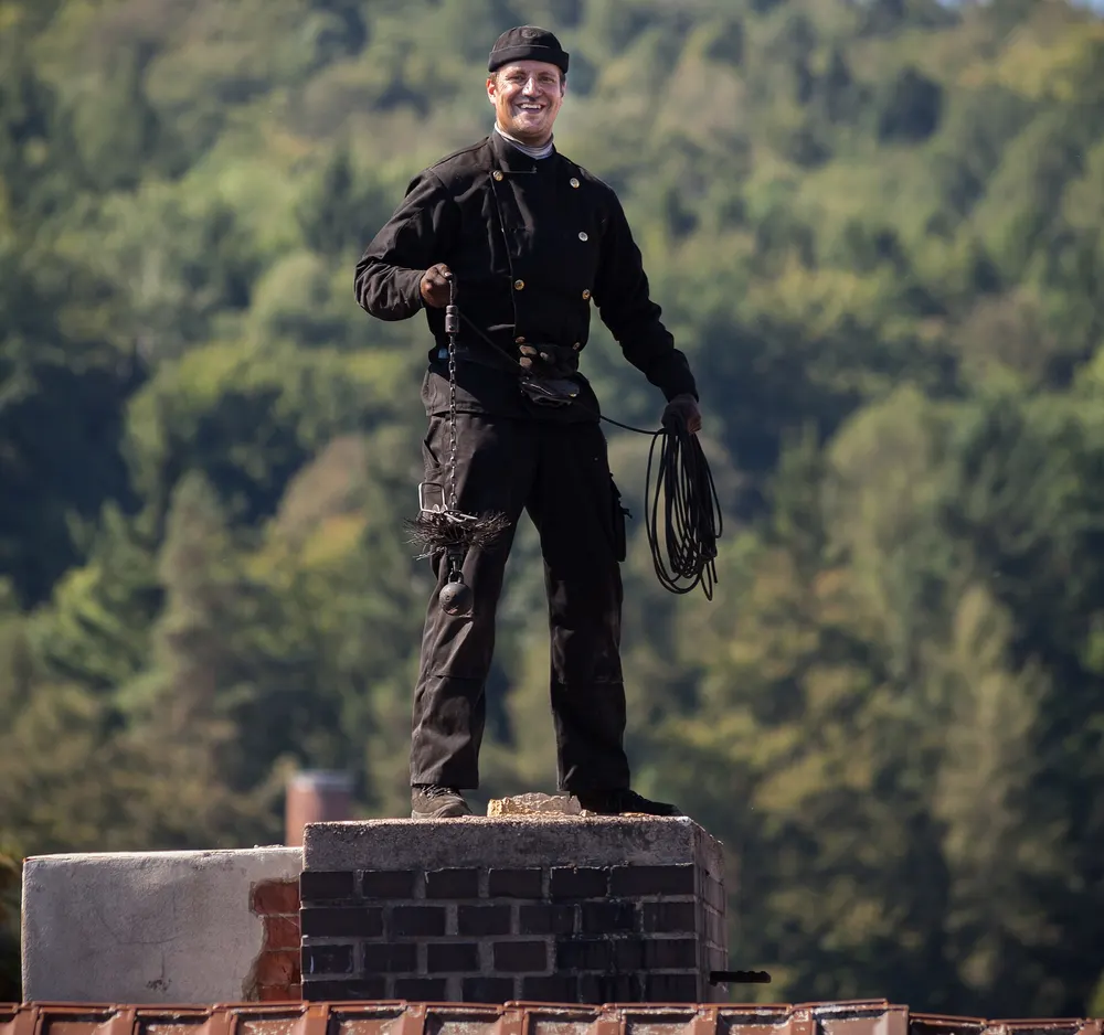 A chimney sweep in black work clothes standing on top of a brick chimney on a tiled roof, holding a coiled sweeping line, with wooded hills behind