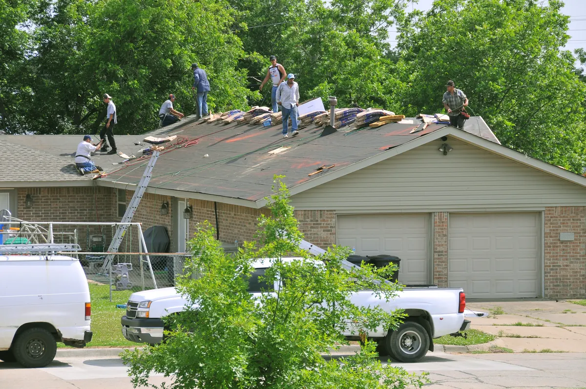 A roofing crew on the roof of a one-story brick house, with bundles of new shingles stacked on bare felt underlayment, a ladder against the eave and work trucks parked in front