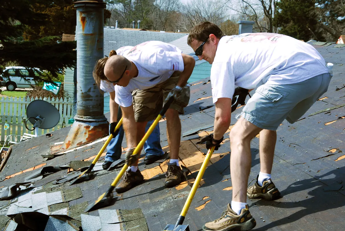 Three people with long-handled roofing shovels stripping old asphalt shingles off a sloped roof, exposing the black felt underlayment and patches of wood decking beside a metal vent pipe