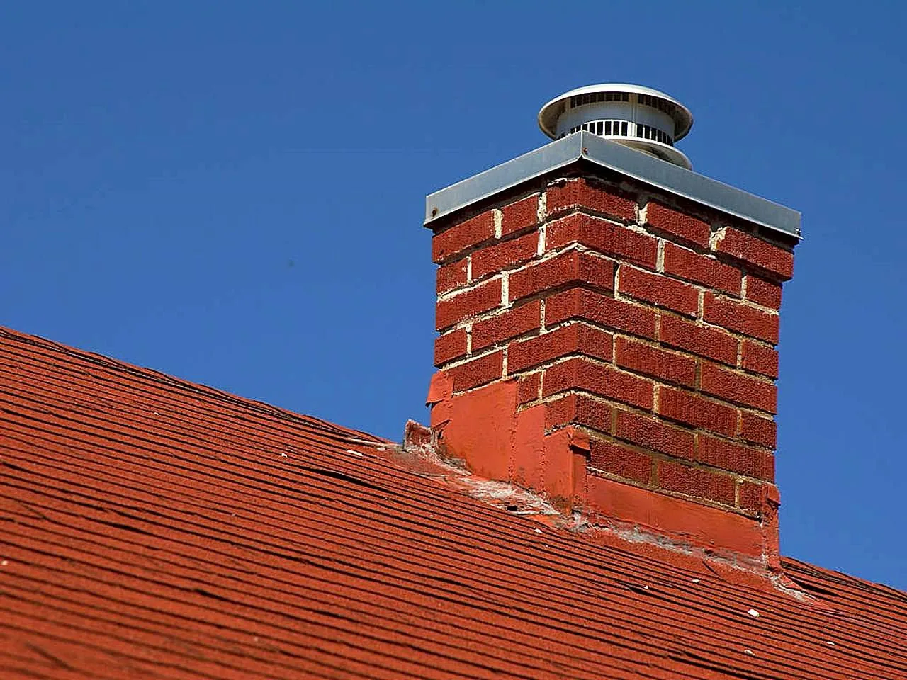 A red brick chimney with a metal rain cap rising through a red asphalt shingle roof against a clear blue sky, with flashing around its base