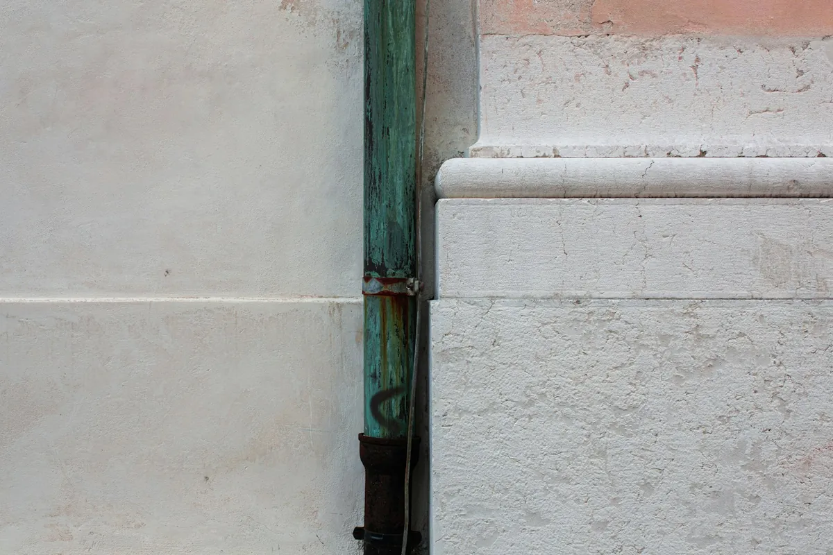 A downspout weathered to streaky blue-green patina running down a pale stone wall beside a carved stone base