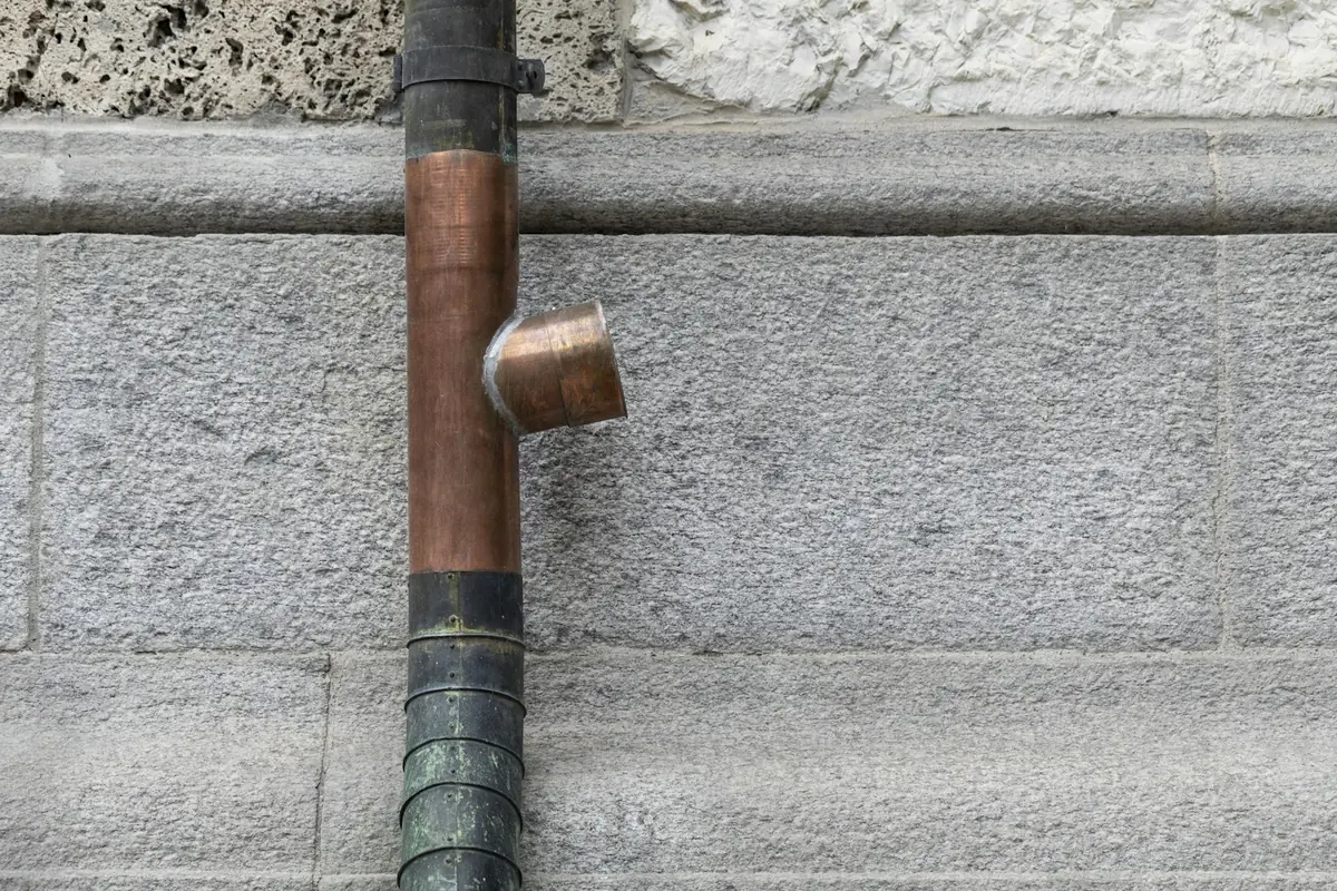 A round copper downspout on a granite wall with a new, bright copper section and a short outlet spliced in between older sections that have darkened and started to turn green