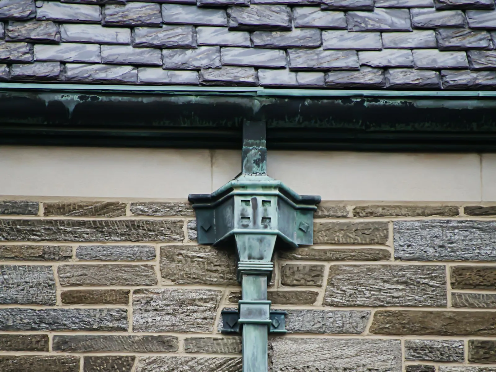 An old copper gutter under a slate roof feeding a decorative copper leader head and downspout, all weathered to a blue-green patina, on a stone building