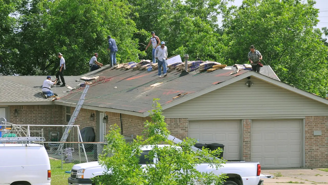 A roofing crew on a one-story brick house stripping the old shingles down to the felt, with bundles of new shingles stacked along the ridge and a vent pipe sticking up through the roof