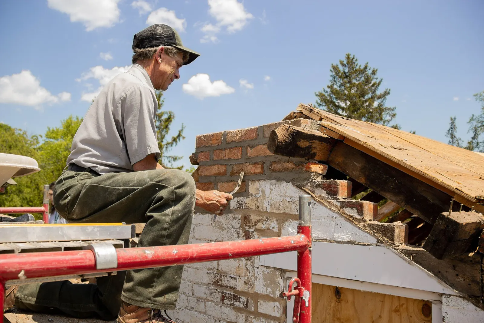 A mason on scaffolding laying new brick and mortar to rebuild a chimney at the gable end of an old house, with the roof boards open beside him