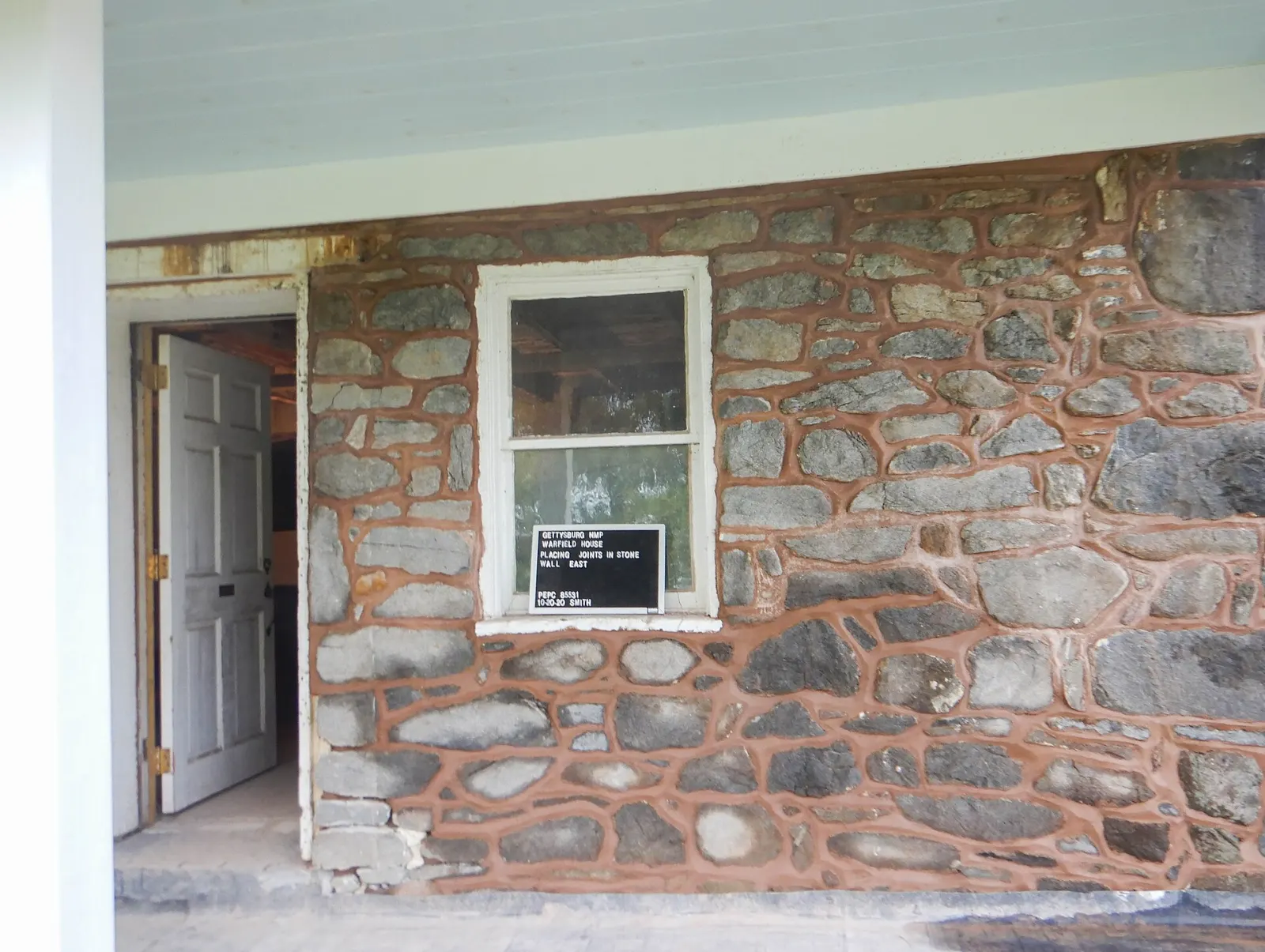 A stone wall of an old house with freshly repointed mortar joints in a reddish brown color around gray stones, next to a window and an open door