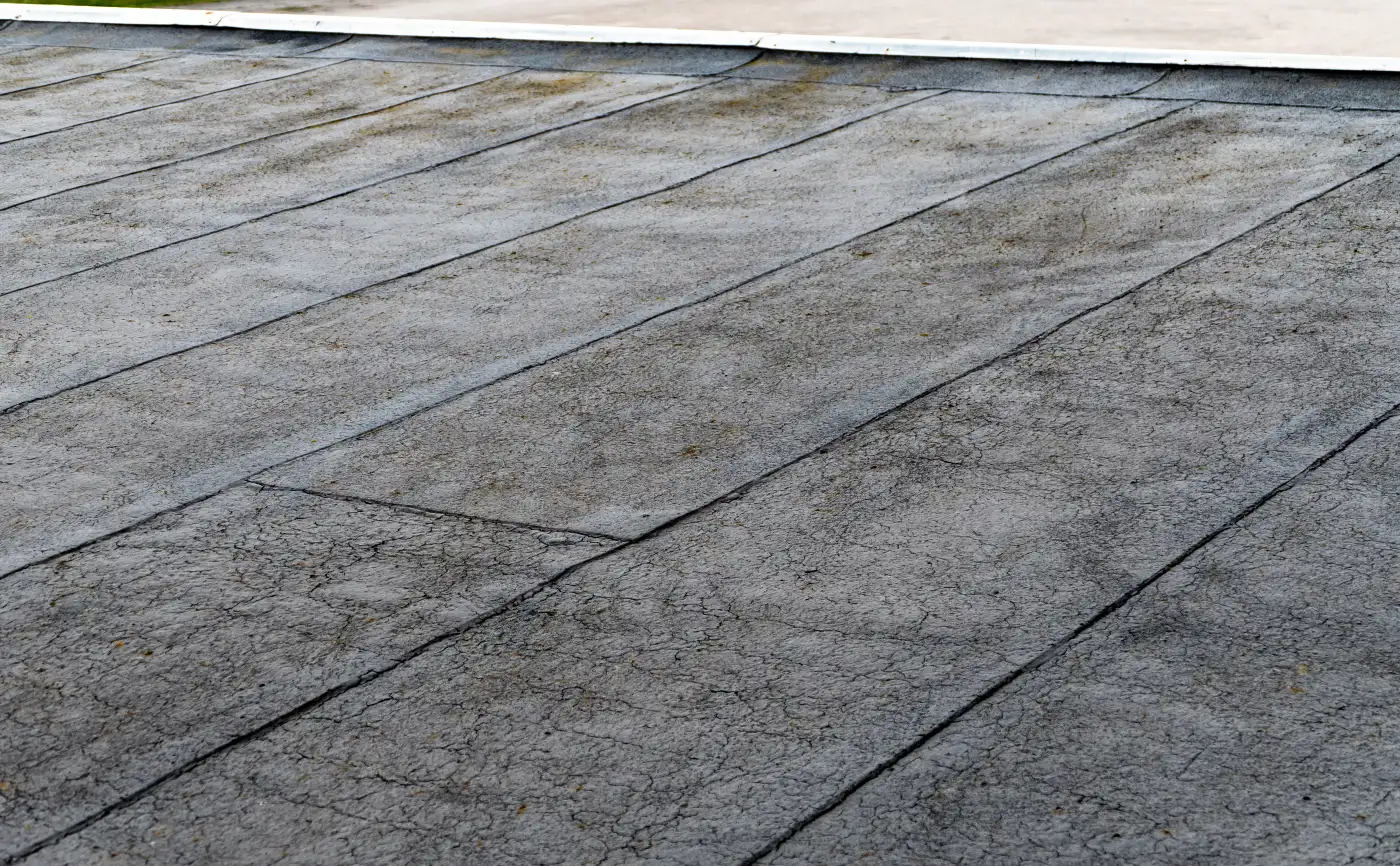 An aged bitumen flat roof with long overlapping seams running across it and surface cracking between the laps