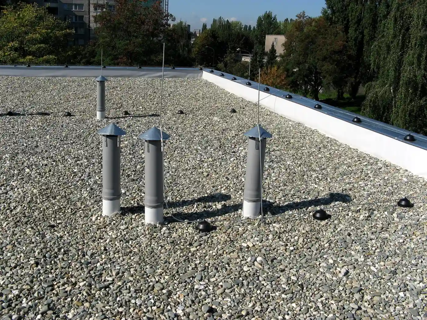 A gravel-surfaced flat roof with three capped vent pipes and lightning protection wires running across it